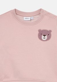Felpa per bambini di colore rosa chiaro con scollo rotondo, bottoni a pressione sulla spalla e una piccola toppa di un orsetto sul petto.