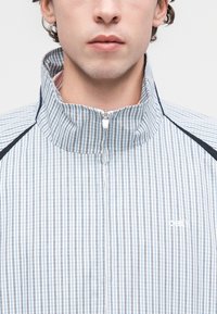 Ελαφρύ τζάκετ με φερμουάρ σε μπλε και λευκές κάθετες ρίγες, με ψηλό γιακά, διακριτικό λογότυπο και αντιθετικά μαύρα στοιχεία στους ώμους.