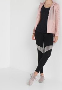 Jasnoróżowa kurtka z kapturem, z białym logo, zestawiona z czarnym topem na ramiączkach i czarnymi legginsami z srebrnymi akcentami. Różowe buty sportowe.