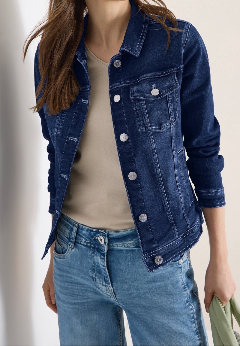Femme portant une veste en jean bleu foncé sur une chemise beige et un jean bleu clair, tenant un tissu vert plié dans la main droite.
