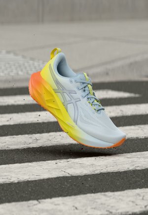 Light blue running shoe with yellow and orange sole, mid-air above black and white striped crosswalk, with gravel falling below.