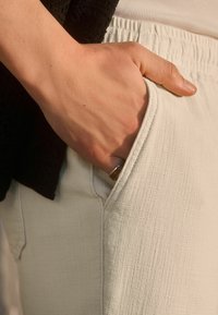 Pantalons blancs texturés avec une poche latérale, main reposant à l'intérieur. Tissu lisse avec des lignes subtiles, complété par un haut sombre. Bague en argent visible.