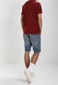 En person som bär en röd t-shirt, blå denimshorts, vita strumpor och vita sneakers står med ryggen mot betraktaren.