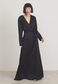 Μαύρο μακρυμάνικο φόρεμα ζώνης με βαθύ V-ντεκολτέ, ζώνη στη μέση, ρευστή σιλουέτα και ομαλή υφή, φτάνοντας μέχρι το πάτωμα.