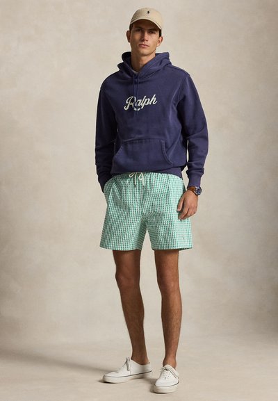 Noor mees, kes kannab beežikat nokamütsi, sodiaagisinu kapuutsi "Ralph" tekstiga, rohelist ruudulist lühikest pükst, valgeid tossusid ja käekella, seisab ühevärvilise tausta ees.