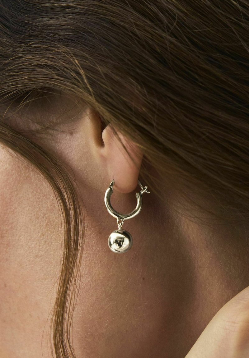 Gros plan sur l'oreille d'une personne aux cheveux bruns portant une boucle d'oreille argentée brillante en forme d'anneau, avec un pendentif sphérique suspendu.