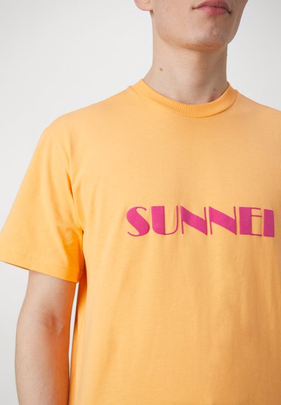 Kortermet t-skjorte med rund hals i lys gul bomull, med stor rosa "SUNNEI" tekst trykket over brystet.