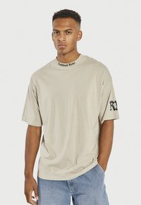 Beżowy t-shirt z krótkim rękawem o luźnym kroju. Posiada czarne logo nadrukowane przy dekolcie oraz czarny akcent na rękawie. Gładka tekstura.