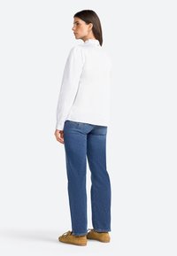 Chemise blanche à manches longues avec un col volanté, associée à un jean en denim bleu et des chaussures beige. La chemise a une coupe ample et des détails de boutons à l'arrière.