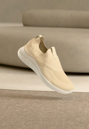Zapatilla de punto beige sin cordones con suela blanca flotando sobre una alfombra beige texturizada, con un sofá beige al fondo.
