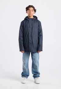 Navy waterproof jacket with a hood, front snap closure, two side pockets, and a smooth texture, worn with light blue jeans and white sneakers.