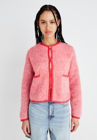 Adīts jaciņa ar sarkanu un rozā svītru rakstu, apaļu kakla izgriezumu, priekšējo pogu aizdari un divām kabatām. Mīksta tekstūra.