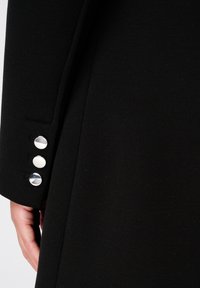 Zwarte jas van gestructureerde stof; voorzien van een verzorgd ontwerp, gladde zilveren knopen en een aansluitende mouwlijn met een nette afwerking.