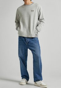 Felpa grigia con scollatura rotonda e un piccolo logo sul petto, abbinata a jeans blu larghi e sneakers bianche.