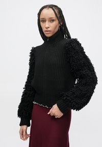 Černý svetr s vysokým límcem, texturovaným pleteným tělem a plyšovými třásněmi na rukávech. V kombinaci s bordó sukní s vlnitým okrajem.