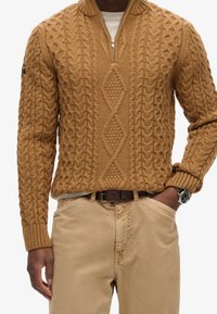 Brauner Pullover mit grobem Strick, hohem Kragen und Reißverschluss. Robuste Textur und Rautenmuster. Kombiniert mit beigen Hosen.