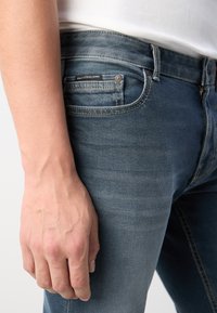 Kék farmernadrág, slim-fit szabással, elhasználódott textúrával, finom varrással és hátsó zseb címkével.