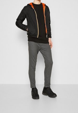 Persona che indossa una giacca nera con fodera arancione, camicia nera, pantaloni grigi e scarpe da ginnastica nere, in piedi contro uno sfondo chiaro e uniforme.