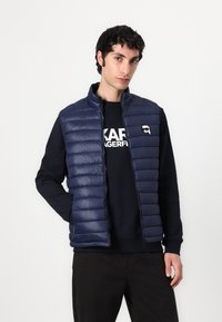 Marineblauwe puffer vest, gequilt ontwerp met hoge kraag en rits aan de voorkant. Daaronder een zwarte sweatshirt met witte letters.