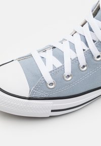 Light blue canvas sneaker with white laces and eyelets, featuring a black rubber sole and a white toe cap. Textured surface with stitching details.