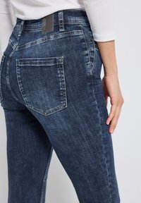 Jean en denim bleu foncé avec une coupe ajustée, doté de poches arrière et de détails de décoloration subtils. Le matériau présente une texture lisse.