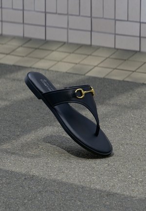 Sandale en cuir noir avec lanière arrière et détail mors de cheval doré, sur un sol en asphalte et un mur carrelé en arrière-plan.