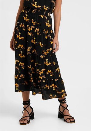 Femme portant une jupe midi noire à imprimé floral jaune et des sandales noires à talons blocs avec lanières, debout devant un fond uni.