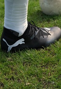 Scarpa da calcio nera con tomaia texturizzata, caratterizzata da un logo Puma bianco, lacci, indossata con calze bianche su un campo d'erba. Palla da calcio sullo sfondo.