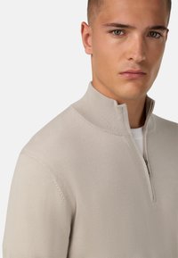 Šviesiai bežinis vilnos megztinis su pusiau užtraukiamu apykakle, raukšlėto audinio ir prigludusio dizaino. Lygus audinys ir minimaliai matomi siuvimo detalės.