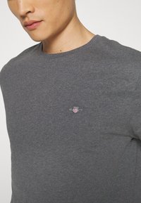 Nahaufnahme einer Person, die ein anthrazitgraues Crewneck-Shirt mit einem kleinen, gestickten GANT-Logo auf der Brust trägt.