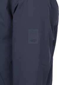 Navyblauwe jas mouw met een rechthoekige rubberen patch logo met een schild, "T," en de tekst "The Swedish Outdoor Brand Est. 1951."