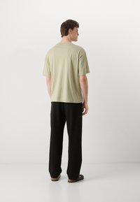 Lys olivenfarvet oversized t-shirt med korte ærmer; parret med sorte vidde bukser og sorte sandaler, set fra ryggen.