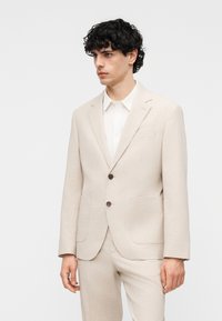 Νεαρός με σκούρα σγουρά μαλλιά, φορώντας μπεζ σακάκι και ταιριαστό παντελόνι πάνω από λευκό πουκάμισο με γιακά, στέκεται μπροστά σε ένα απλό φόντο.