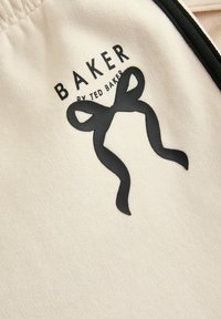 Un tissu crème présente un logo noir et brillant avec le texte "BAKER" et un design de nœud décoratif, souligné pour un accent visuel.