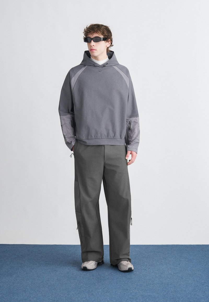 Grå hættetrøje med teksturerede ærmer, kombineret med brede grå bukser med lynlåse. Modellen bærer mørke, elegante solbriller og sneakers.