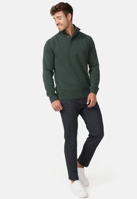 Pull en maille vert foncé avec un col haut, trois boutons et des poignets côtelés, associé à un pantalon slim gris foncé et des baskets blanches.