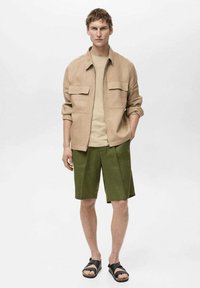 Beige linnedjakke med frontlommer, beige t-shirt, grønne plisserede shorts og sorte sandaler, der skaber et lagdelt, afslappet outfit.