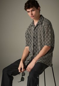 Camicia a maniche corte con motivo bianco e nero e bottoni, indossata con pantaloni neri. Il modello tiene degli occhiali da sole, seduto su uno sgabello contro uno sfondo neutro.