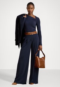 Marineblauwe langemouwen top, wijde broek met een bruine riem en een bruine handtas met krokodillenstructuur. Model houdt een donkere jas vast.