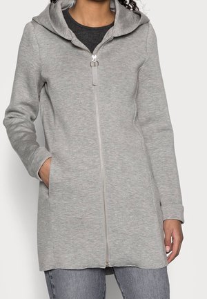 Femme portant un sweat à capuche long gris clair zippé avec des poches, associé à un jean gris et un haut foncé en dessous.