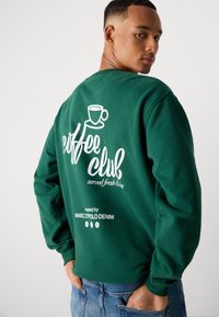 Man in een groene sweatshirt met de tekst "Coffee Club geserveerd vers & heet, gebrand door Marc O'Polo Denim" in het wit en een koffieblik afbeelding op de achterkant.