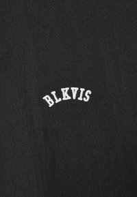 Melna auduma fona ir izlieka balts izšūta logo ar uzrakstu "BLKVIS", kas izceļas pret tumšo fonu.