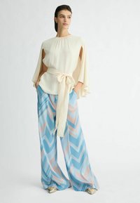 Cremefarbene Bluse mit Capeärmeln und gebundenem Taillengürtel, kombiniert mit weitgeschnittenen Hosen im blau-pink gestreiften Zickzackmuster. Geschmeidiger, leichter Stoff.