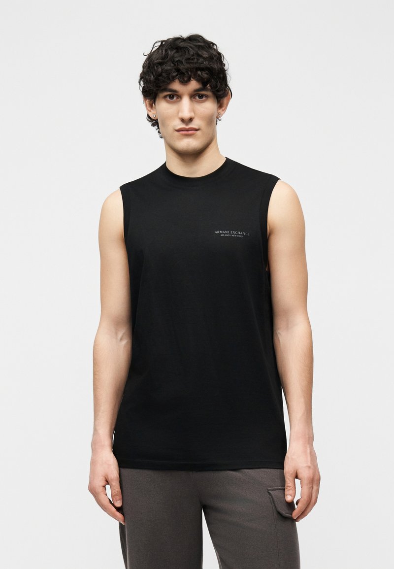 Jeune homme aux cheveux bouclés portant un débardeur noir Armani Exchange et un pantalon gris foncé sur un fond uni.