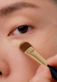 Makeupbørste med et gyllent håndtak påfører kremete concealer under øynene, som forbedrer den jevne hudoverflaten; varm brun øye i fokus.