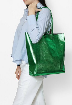 Borsa tote metallica verde con una finitura testurizzata e manici lunghi, portata da una persona. La borsa presenta una forma rettangolare ampia.