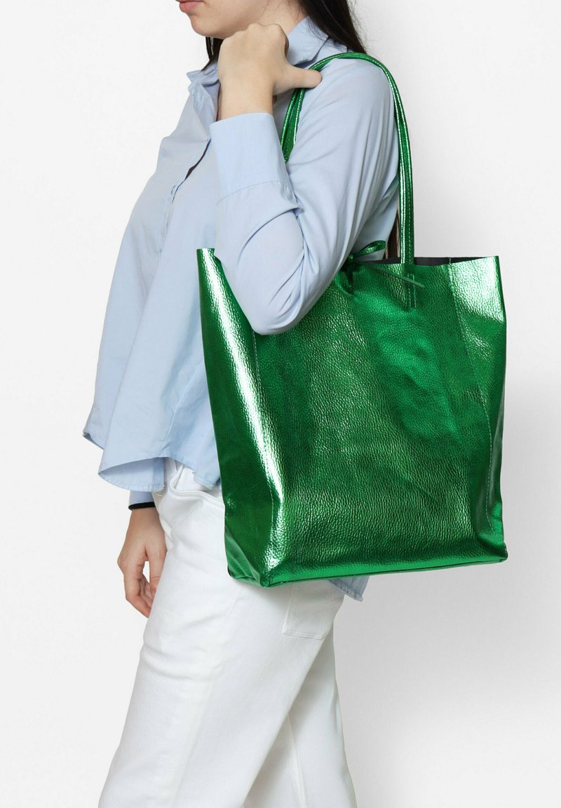 Sac fourre-tout métallique vert avec une finition texturée et de longues poignées, porté par une personne. Le sac présente une forme rectangulaire spacieuse.