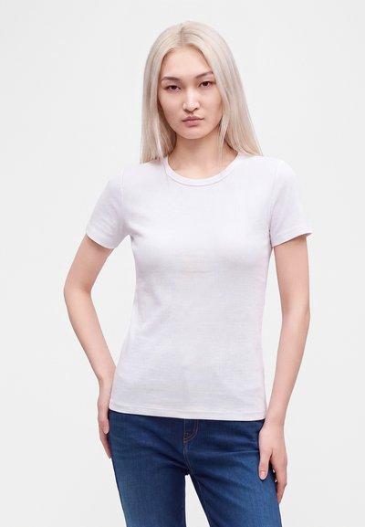 Jeune femme aux longs cheveux blonds et raides, portant un t-shirt blanc uni à manches courtes et un jean bleu, debout avec une main sur la hanche.