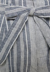 Tessuto a righe con linee verticali blu navy e bianche; presenta una cintura in tessuto e una piccola tasca laterale. Materiale texturizzato.