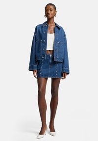 Ensemble en jean sombre composé d'une veste et d'une jupe, au design ample, avec des fermetures à boutons et des accents argentés. Associé à un top blanc et des chaussures.